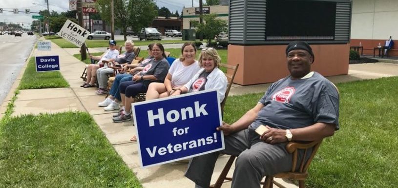 Several people sit on rocking chairs on a sidewalk beside Davis College holding signs that say 'honk for Veterans'.